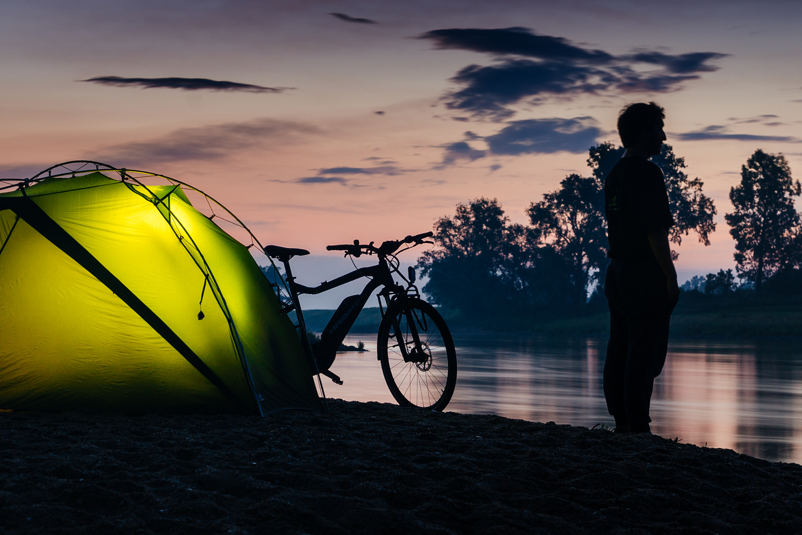 Mann neben beleuchtetem Zelt mit E-Bike am Fluß