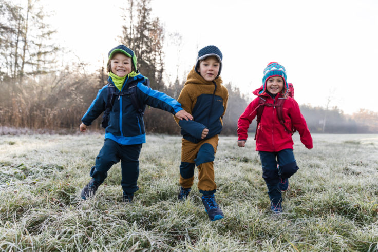 Drei Kinder mit Bekleidung aus Recycling-Materialien springen voller Freude über gefrorene Wiese.